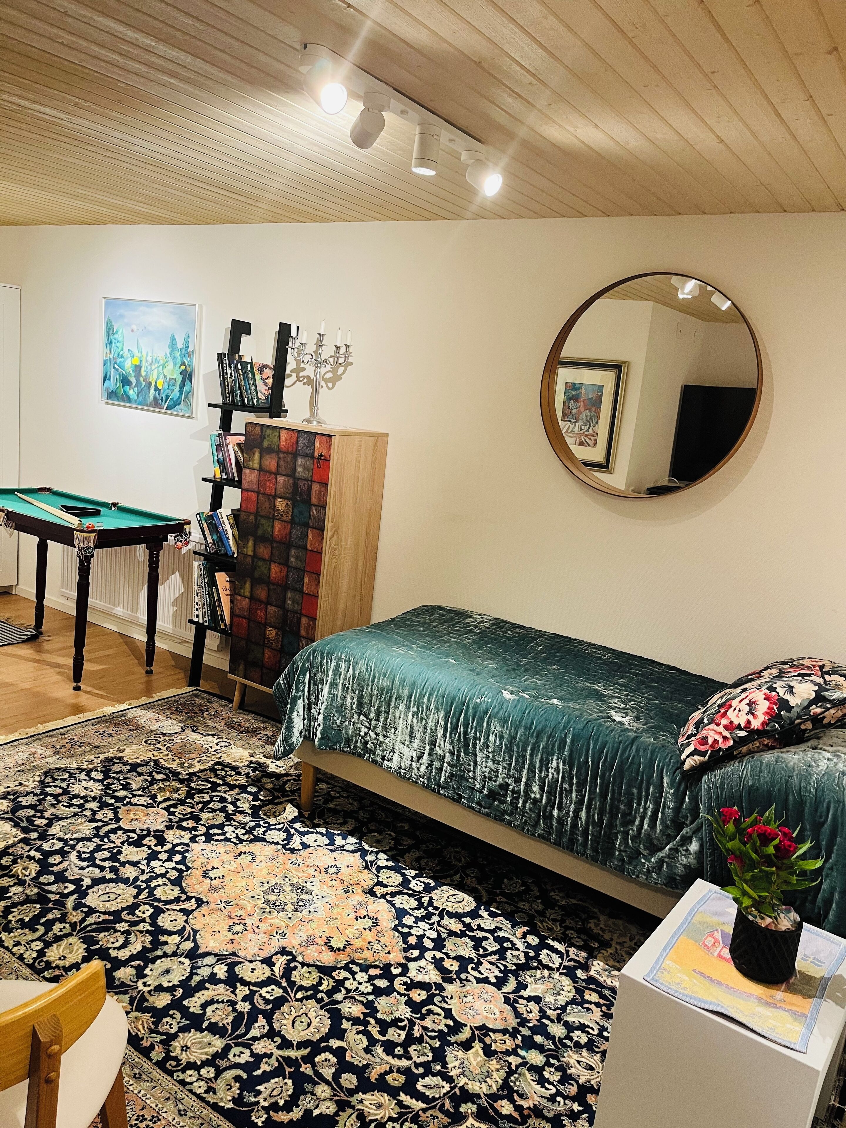 Un dormitorio con cama, escritorio, estantería, espejo y alfombra.