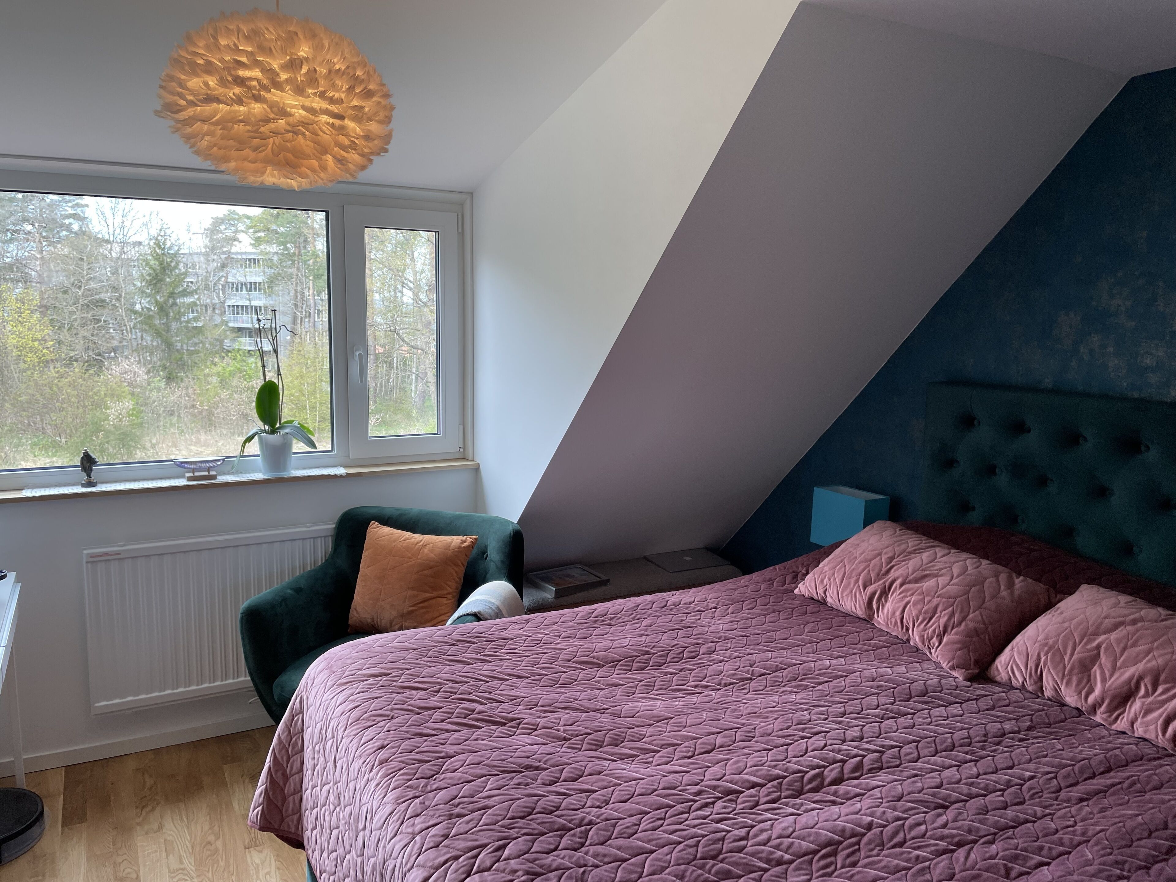 A bedroom with a sloped ceiling, a bed with a pink quilted bedspread, a green armchair, a window with a view of trees, and a unique textured ceiling light.