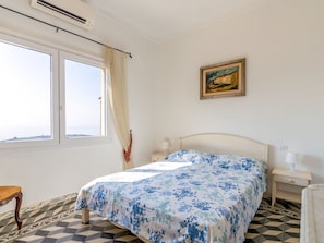 Een slaapkamer met een bed, nachtkastje, lamp, schilderij aan de muur en een raam met gordijnen.