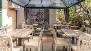 Een terras buiten met houten tafels en stoelen, een glazen dak en een stenen muur op de achtergrond.