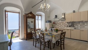 Un coin repas avec une table dressée pour quatre personnes, des chaises en bois, un lustre et une vue sur la mer, visible à travers une arche ouverte.