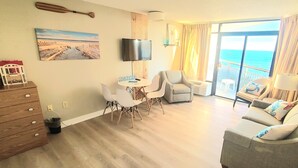 A spacious living room with a flat-screen TV, a painting of a beach, a dining area with a small table and chairs, and a balcony with a view of the ocean. - Ocean View King Suit Heated Pools Myrtle Beach  (Myrtle Beach)