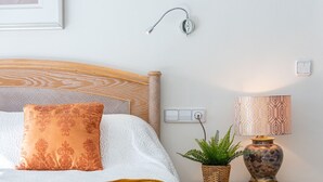 Ein ordentlich eingerichtetes Schlafzimmer mit einem Bett, einer Nachttischlampe, einer Wandleuchte, einer Steckdose und einer Zierpflanze.
