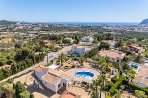 A residential area with a swimming pool, surrounded by houses with terracotta roofs, and a view of the coastline.