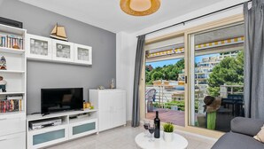 A modern living room with a flat-screen TV, white cabinets, and a balcony with a view of buildings and greenery. - Apartamento Zeus 6 - PlusHolidays (Calpe)