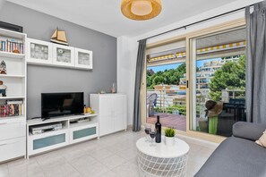 Ein modernes Wohnzimmer mit Flachbildfernseher, weißen Schränken und einem Balkon mit Blick auf Gebäude und Grünflächen.
