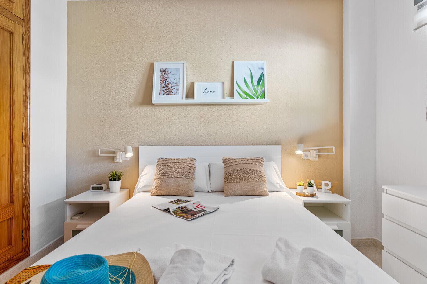A neatly arranged hotel room with a large bed, two bedside tables, a nightstand, a wall-mounted shelf with framed pictures, and a bedside lamp.