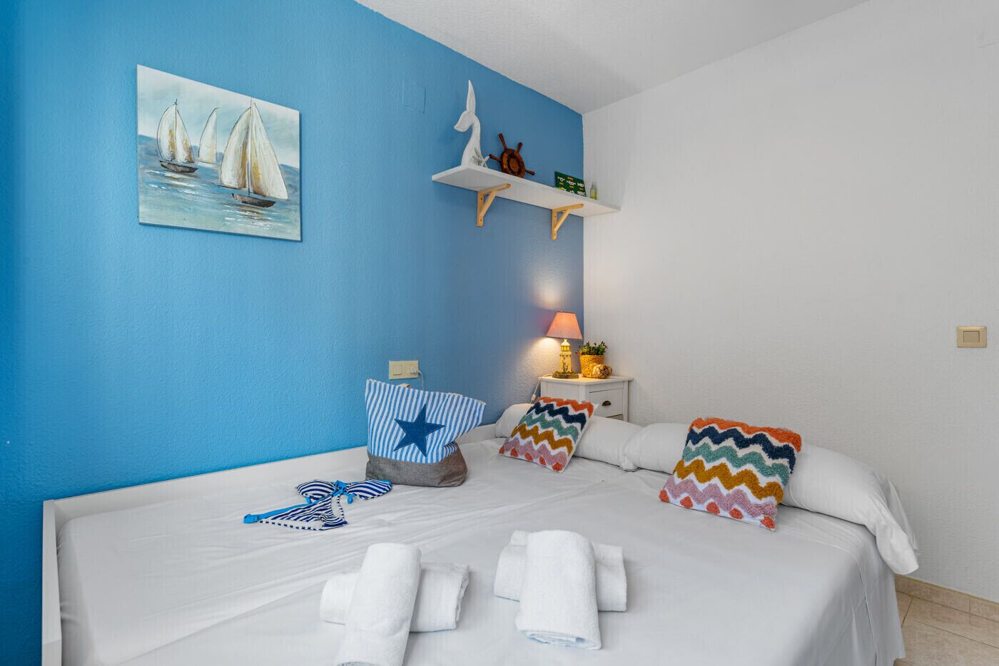 A bedroom with a blue wall, a bed with white bedding, a painting of sailboats, a shelf with a ship's wheel, and a nightstand with a lamp and a plant.