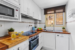 A compact kitchen with white cabinets, a microwave, an oven, and a dishwasher. There are potted plants on the counter, and a window with a view of the outdoors. - Apartamento Zeus 6 - PlusHolidays (Calpe)