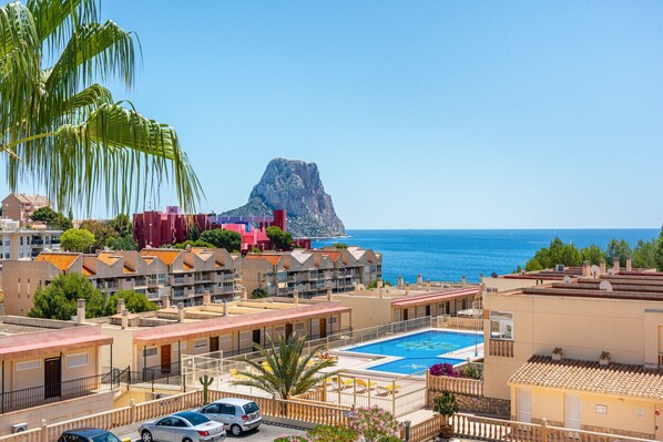 A coastal hotel with a swimming pool, balconies, and a view of the sea and a prominent rock formation.
