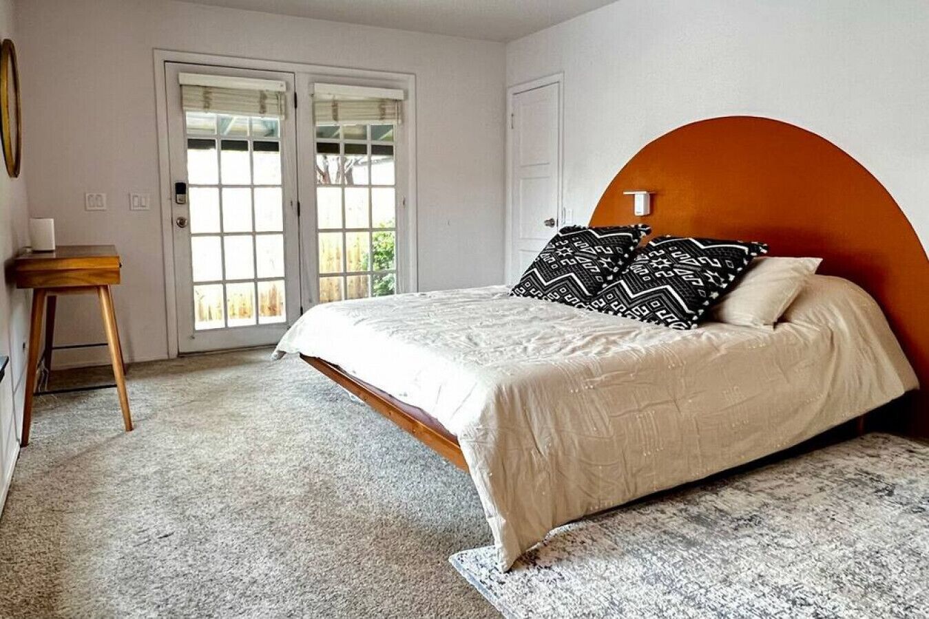 A bedroom with a bed, a wooden nightstand, a window with blinds, and a door.