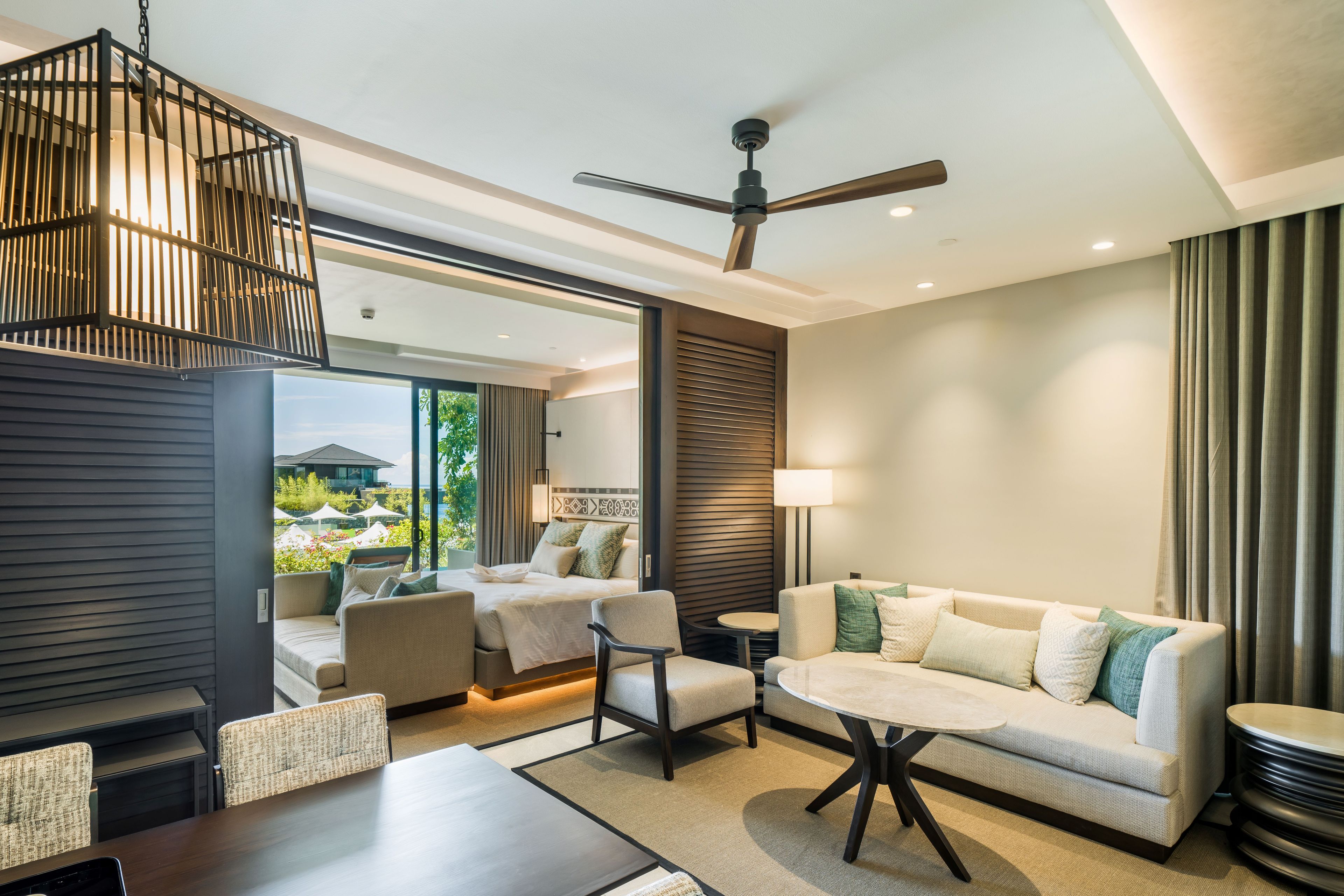 A modern hotel room with a balcony, a ceiling fan, a sofa, a dining table, and a view of the outdoors.