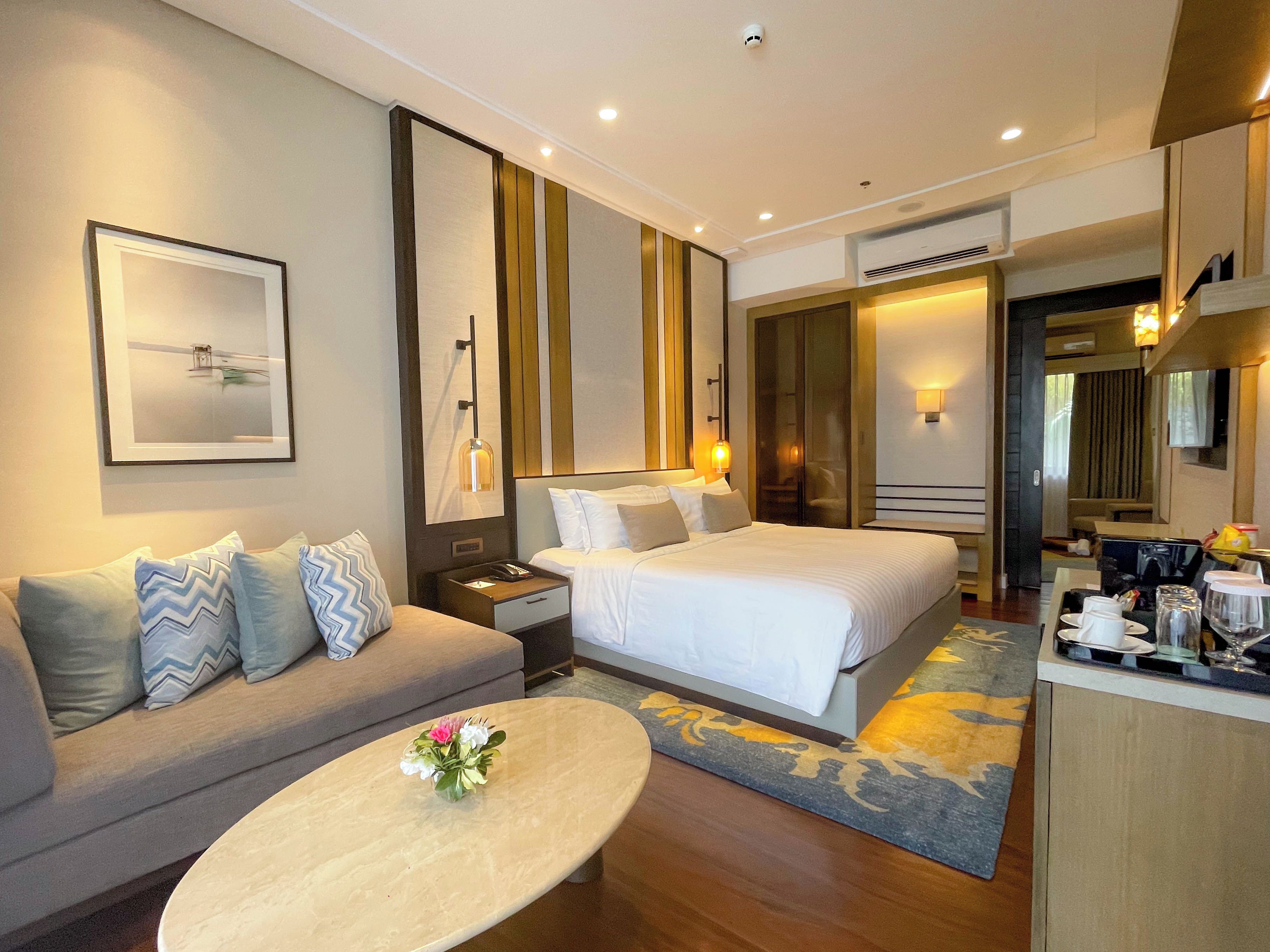 A hotel room with a sofa, two beds, a coffee table with flowers, a framed picture, and a mirror.