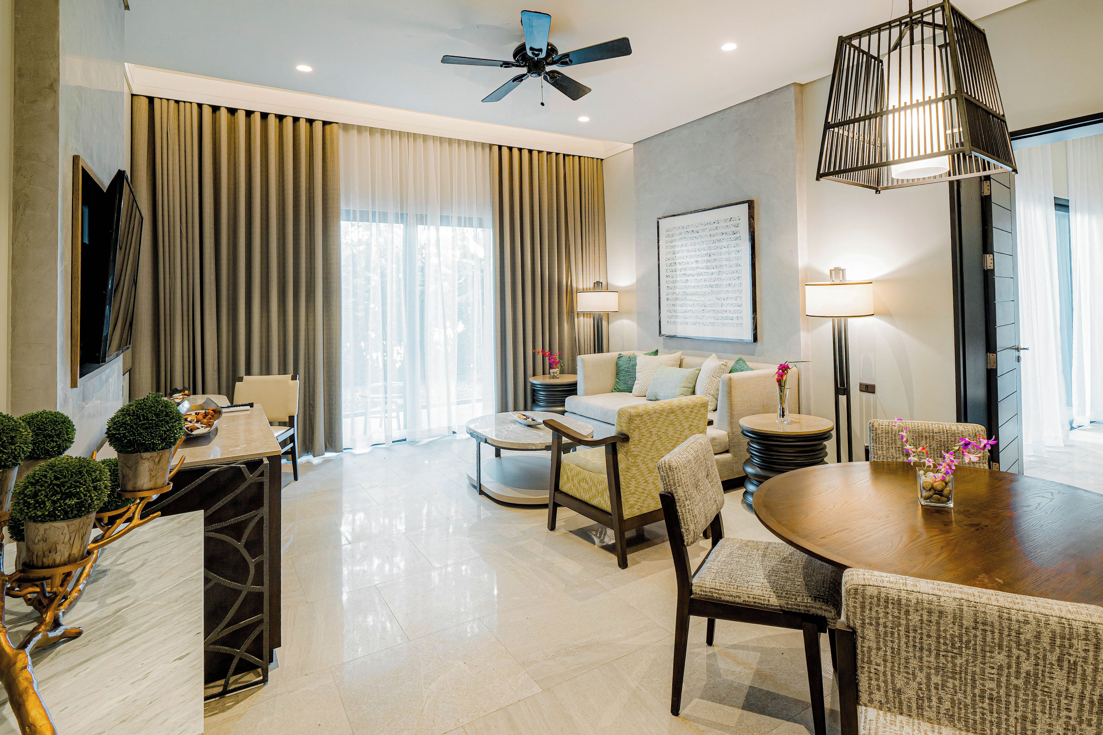 A modern hotel room with a dining area, a sofa set, a ceiling fan, and a large window with curtains.