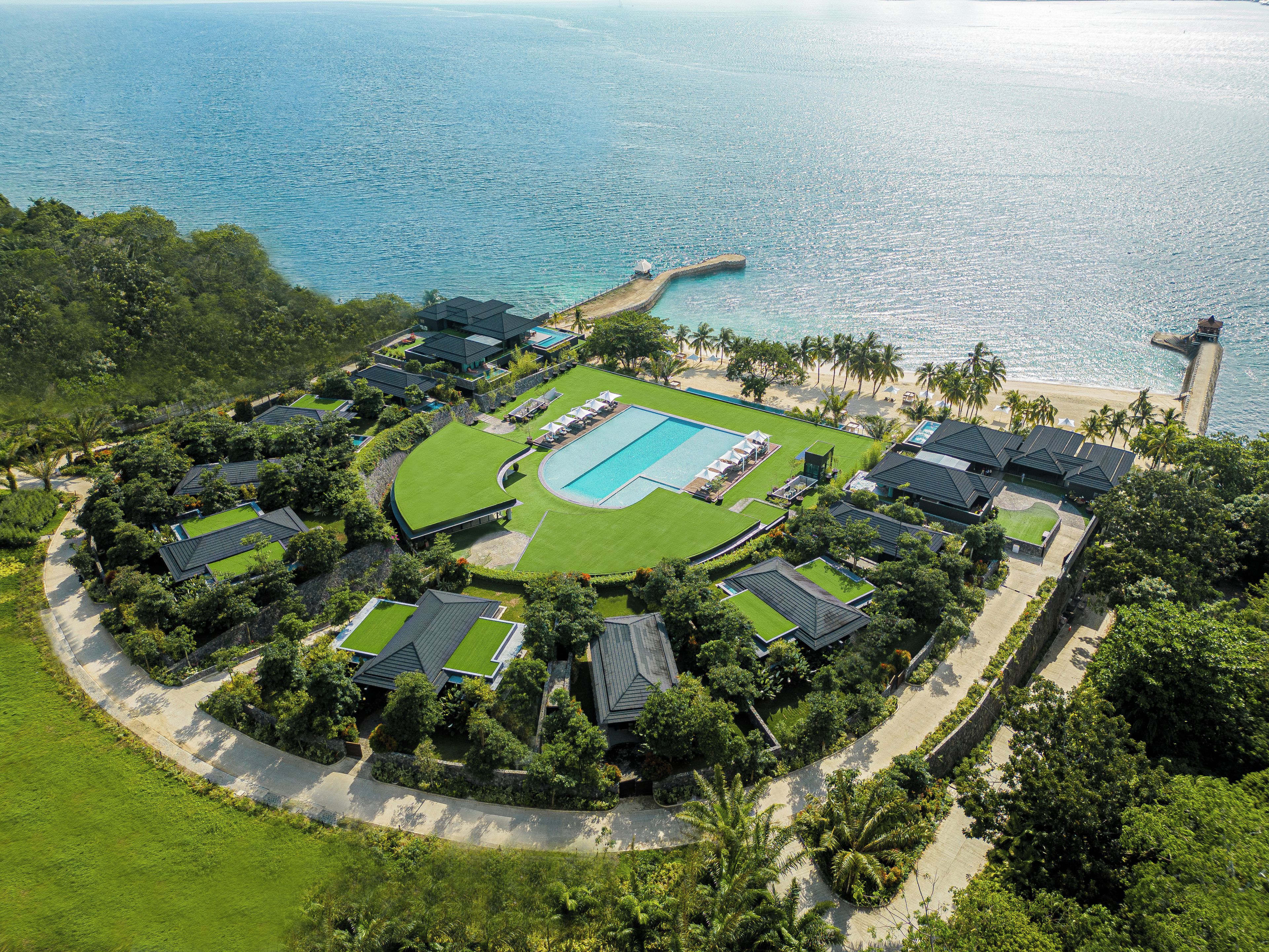 Vista aérea de un complejo turístico con piscina central, varias villas para huéspedes y una playa con palmeras.