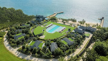 Vista aérea de un complejo turístico con piscina central, varias villas para huéspedes y una playa con palmeras.