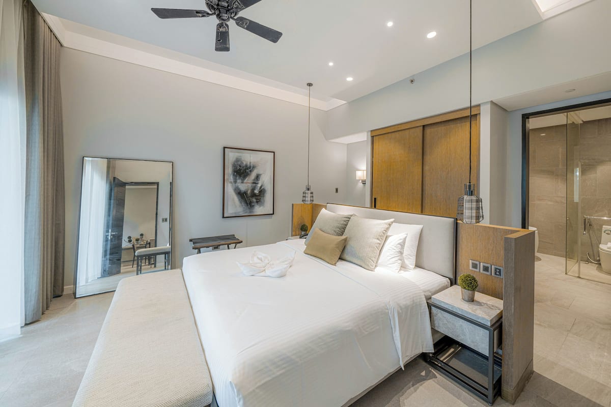 Quarto de hotel moderno com uma cama grande, um ventilador de teto, uma mesa de cabeceira com abajur, um espelho e vista para o banheiro.