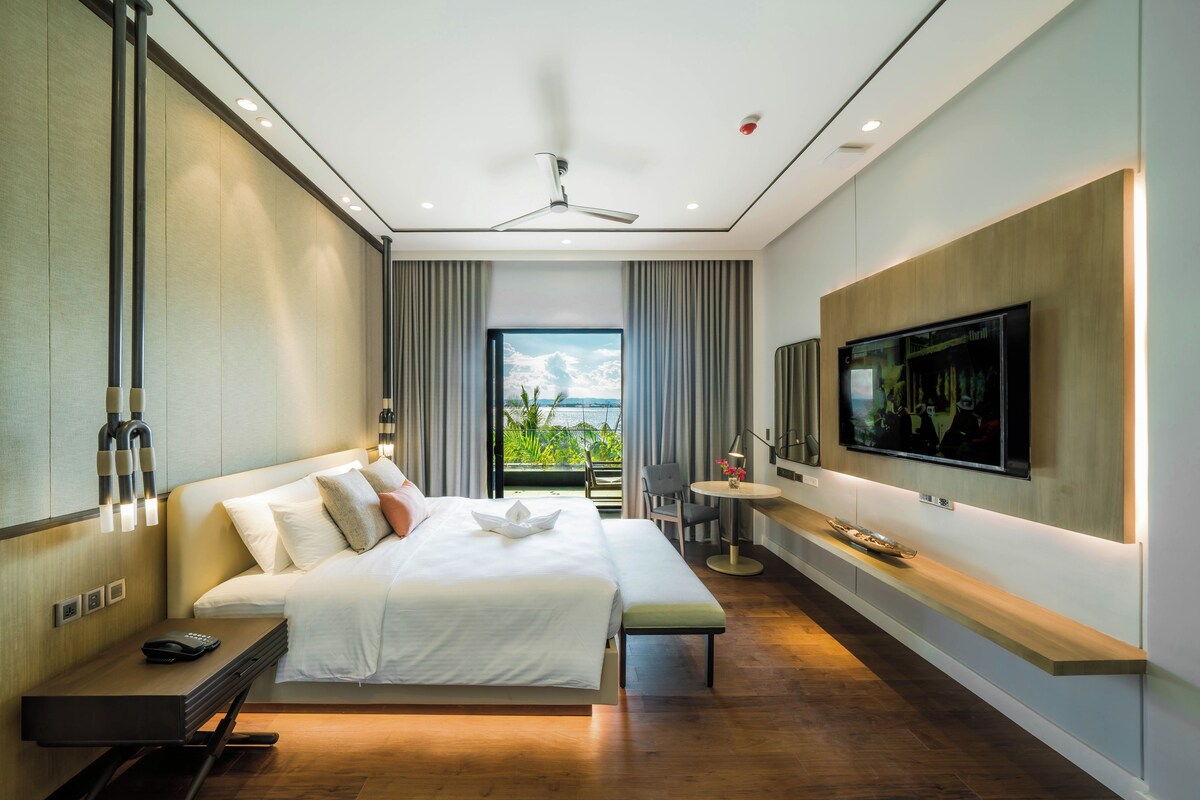 A modern hotel room with a large bed, a desk, a chair, a TV, and a balcony with a view.