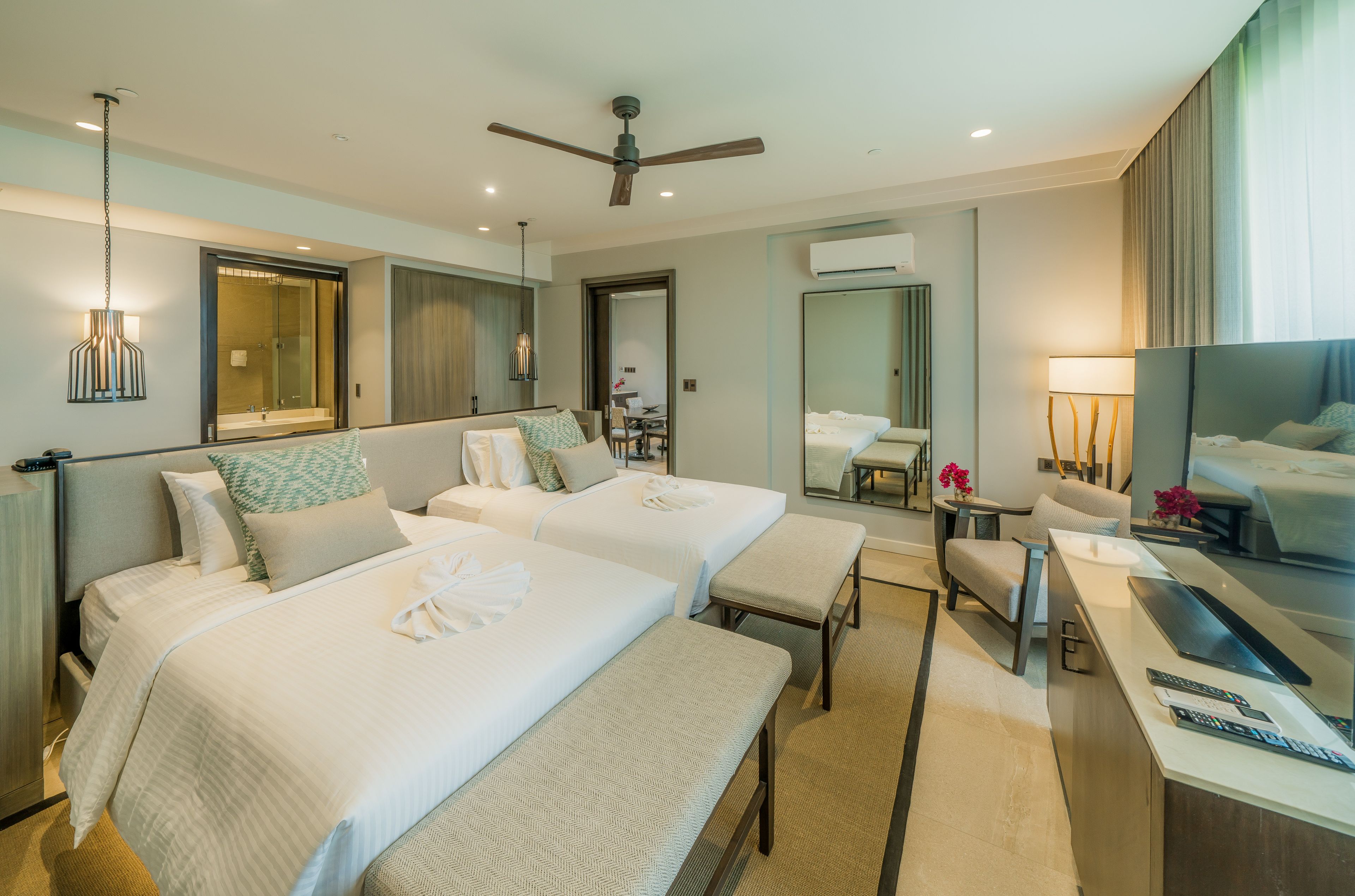A hotel room with two beds, a desk with a TV, a ceiling fan, and a bathroom visible through a glass door.