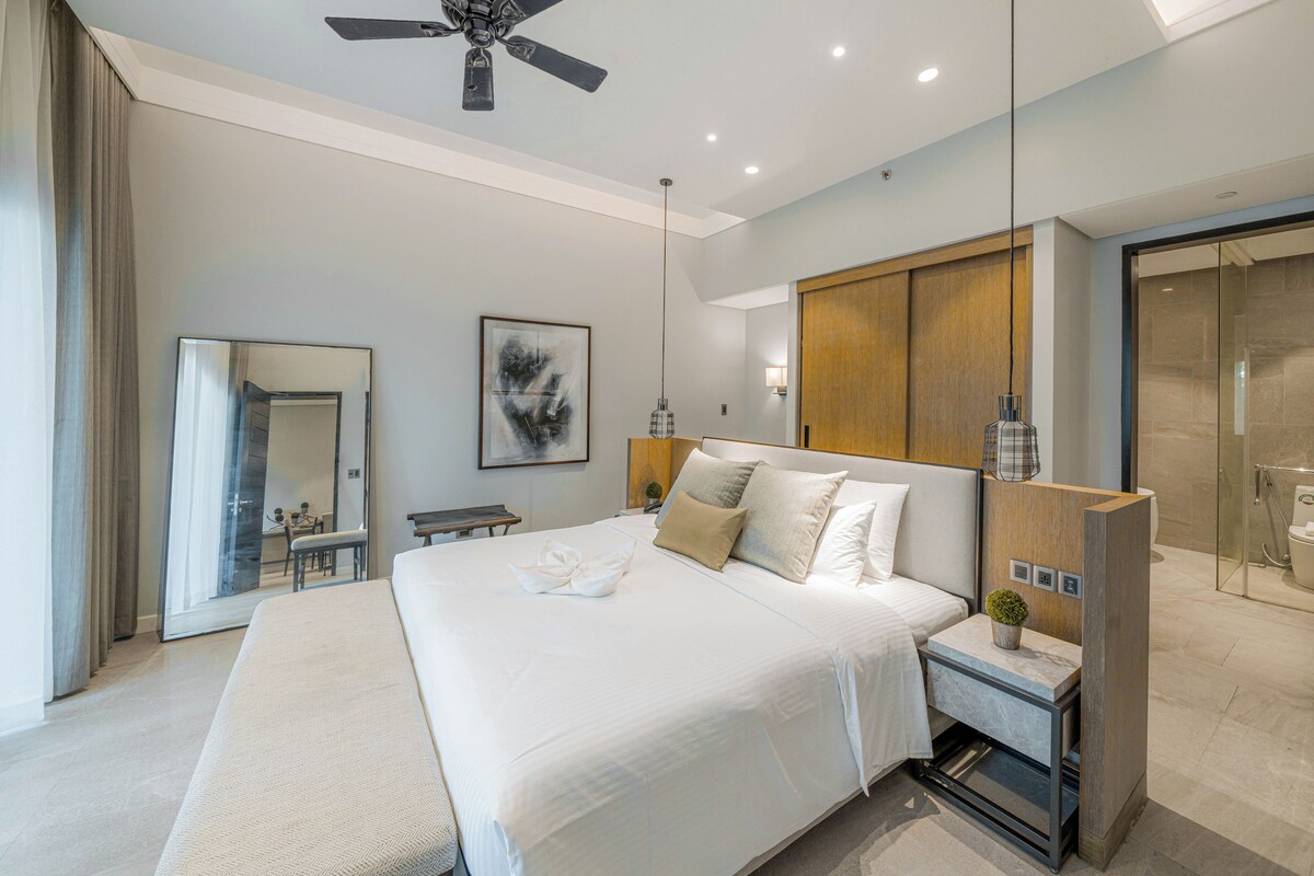 A modern hotel bedroom with a large bed, a ceiling fan, a bedside table with a lamp, a mirror, and a view of the bathroom.