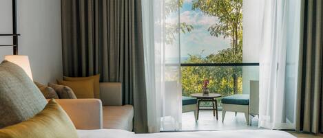 Habitación de hotel con una cama grande, un sofá, un balcón con mesa y sillas, y vistas a zonas verdes.