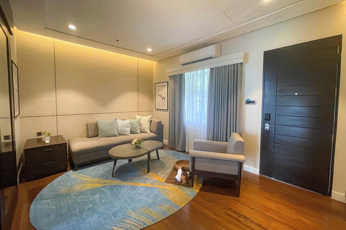 A modern living room with a sofa, a coffee table, and a chair. There is a large window with curtains, a door, and an air conditioning unit on the wall.