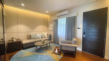 Una sala de estar moderna con un sofá, una mesa de centro y una silla. Hay un ventanal con cortinas, una puerta y un equipo de aire acondicionado en la pared.