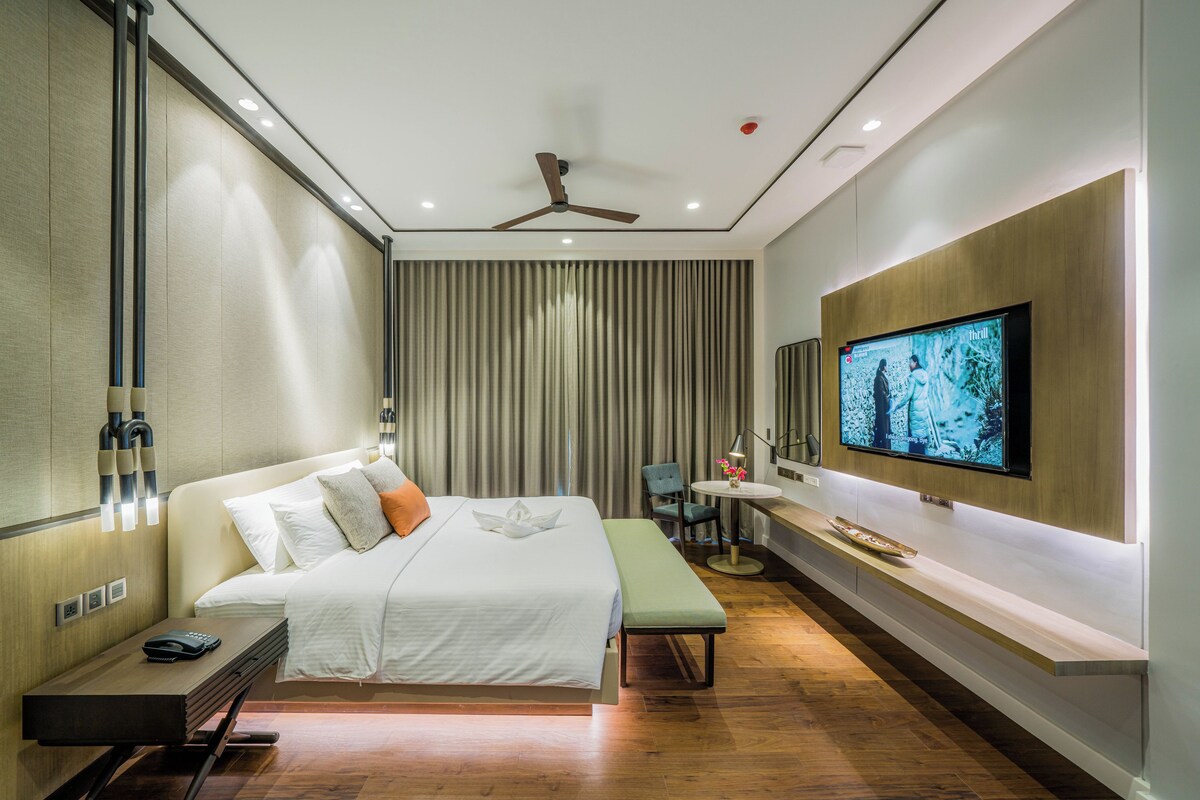 A modern hotel room with a large bed, a desk, a chair, a TV, and a ceiling fan.