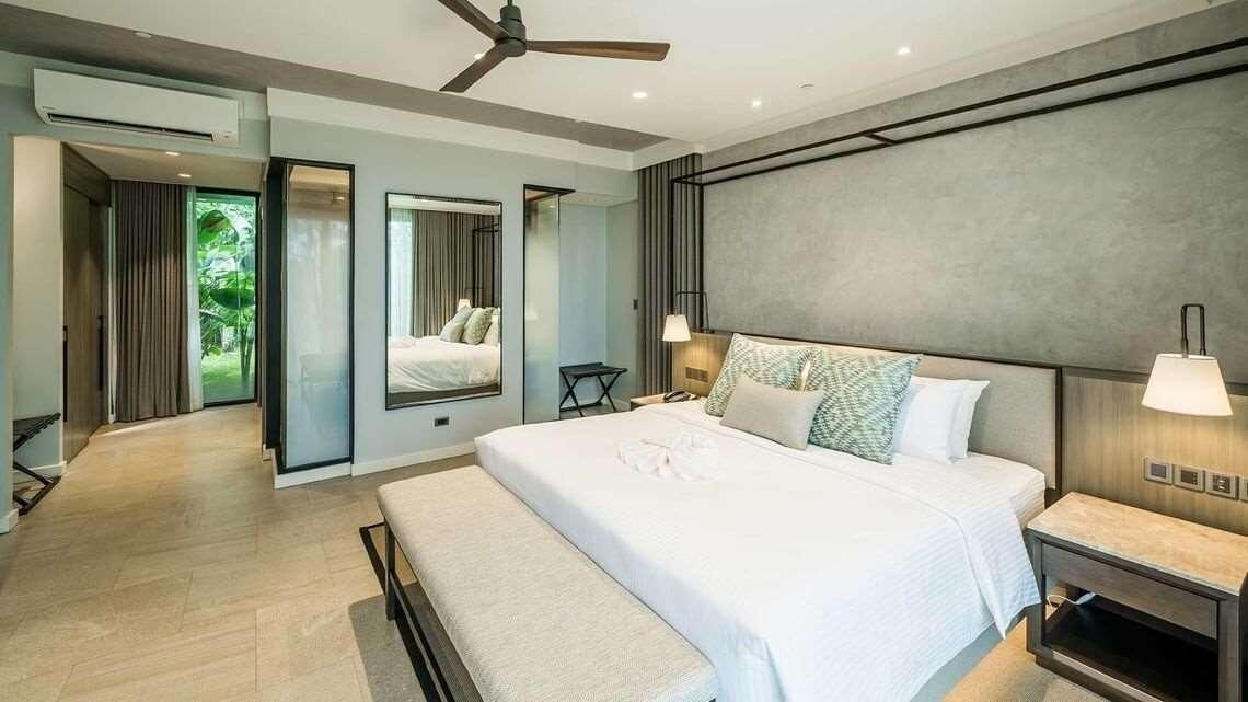 Une chambre moderne avec un grand lit, des tables de chevet, un ventilateur de plafond et une vue sur un jardin, accessible par une porte ouverte.