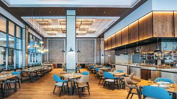 A spacious restaurant with multiple dining tables and chairs, a kitchen area, and large windows allowing natural light.