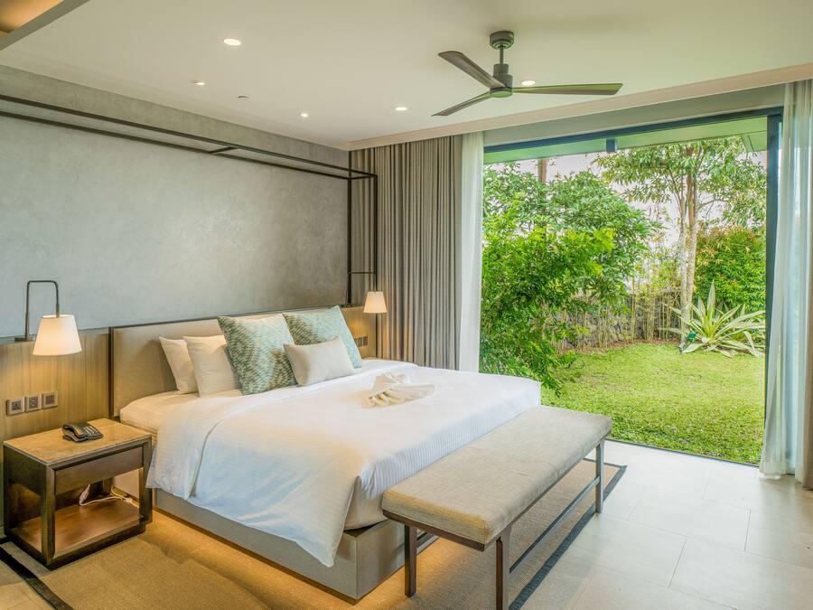A modern hotel room with a large bed, bedside lamps, a nightstand, a bench, and a view of a lush garden through a sliding glass door.