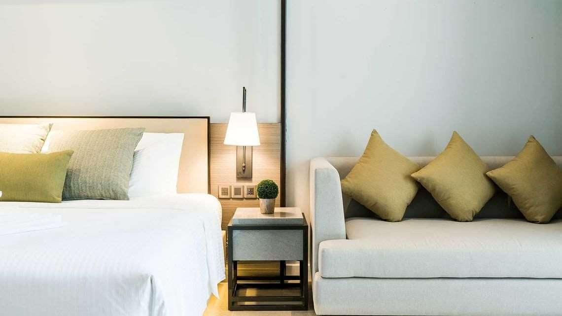 Habitación de hotel con cama, mesita de noche con lámpara, pequeña mesa con planta y sofá con cojines.