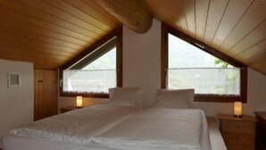 A bedroom with a wooden ceiling, a bed with white linens, two bedside tables with lamps, and windows with curtains. - Rustico Cleo by Interhome (Semione)