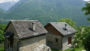 Two stone buildings with sloped roofs, set against a backdrop of lush green mountains. - Rustico Cleo by Interhome (Semione)