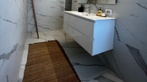 Ett modernt badrum med en vägg av marmor, ett vitt handfat med tillhörande skåp, en spegel och en brun matta.
