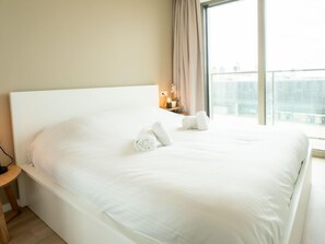 A neatly made bed with white linens and two rolled towels, a bedside table with a lamp, and a large window with a view of buildings.