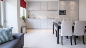 A modern kitchen with white cabinets, a dining area with white chairs, and a grey sofa with a patterned pillow. - Apartment Andrei by Interhome (Blankenberge)