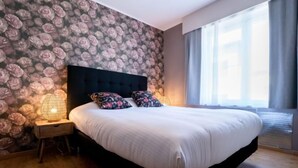 A bedroom with a large bed, floral wallpaper, a nightstand with a lamp, and a window with curtains. - Philemon with parking by Interhome (Blankenberge)