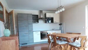 A modern kitchen with white cabinets, a built-in oven, and a dining area with wooden chairs and a table. - Löwenzahn by Interhome (Velden am Wörthersee)