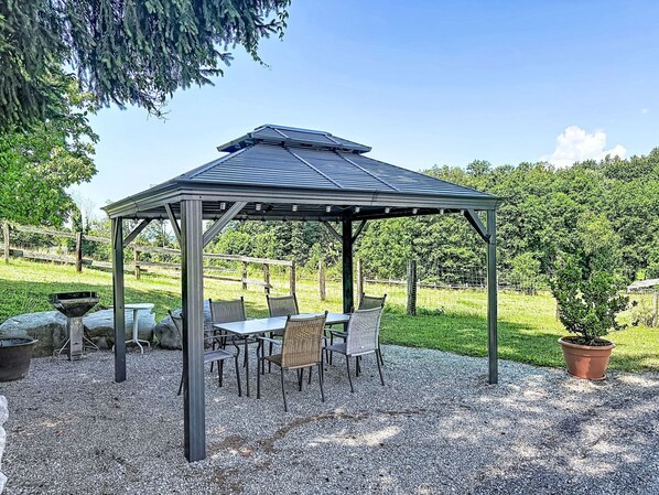 A gazebo with a metal roof and a gravel floor, surrounded by chairs and a table.