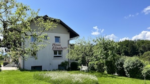 Un edificio llamado "Haus Elisabeth" con fachada blanca, un pequeño jardín con flores blancas y un cielo azul despejado.