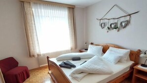 Una cama de madera con ropa de cama blanca, una silla roja, una mesita de noche con una lámpara y una ventana con cortinas translúcidas.