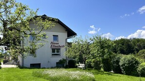 Un edificio llamado "Haus Elisabeth" con fachada blanca, un pequeño jardín con flores blancas y un cielo azul despejado.
