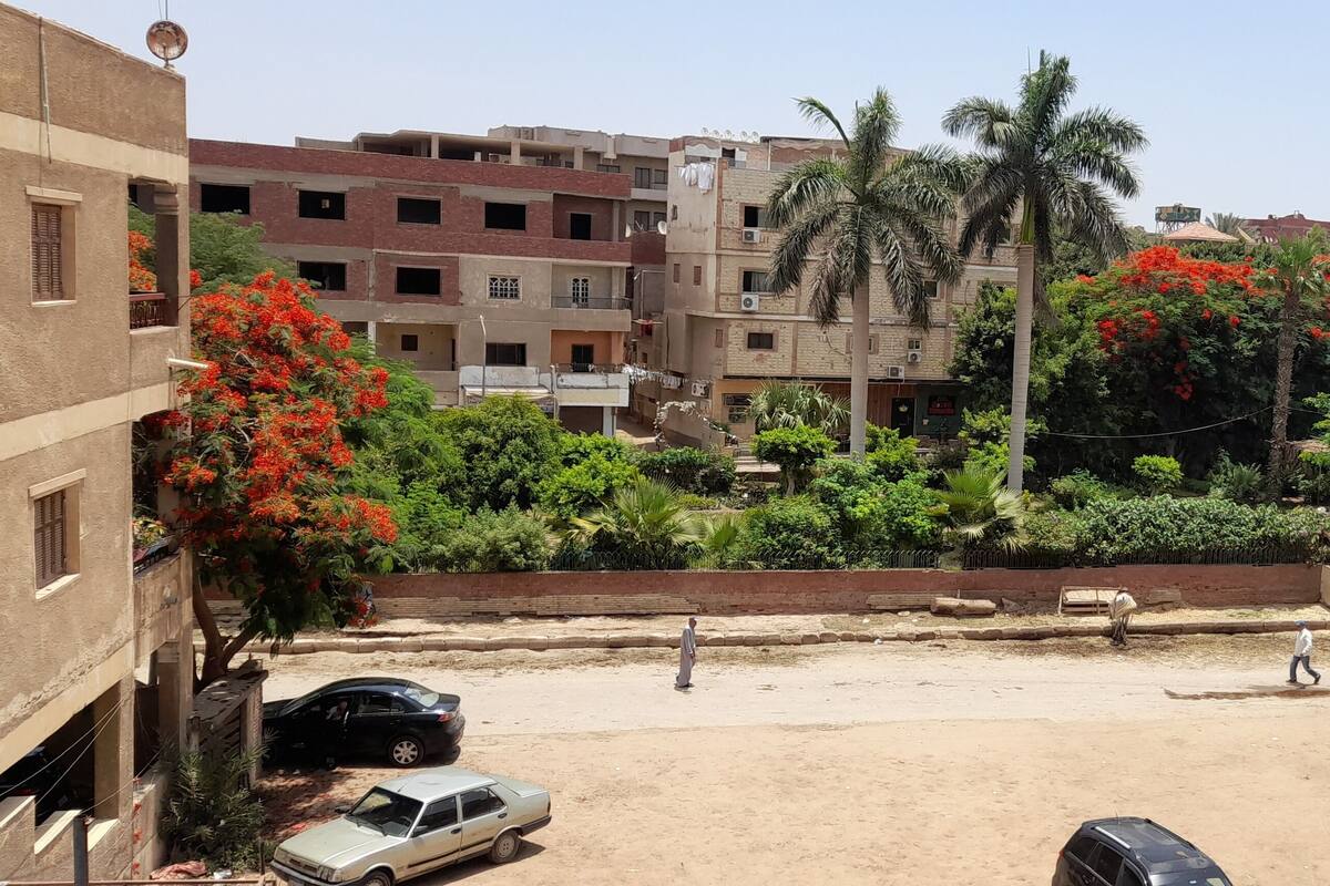 Un quartier résidentiel avec des immeubles à plusieurs étages, une rue sablonneuse et une végétation luxuriante, notamment des arbres à fleurs rouges.
