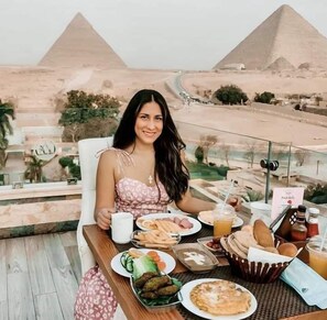 A dining table with food and drinks, a view of pyramids, and a cityscape in the background.