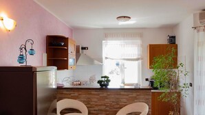 A kitchen with two bar stools, a stone backsplash, wooden cabinets, a potted plant, and a window with curtains. - KATA Two-Bedroom Apartment with Balcony (Rovinj)