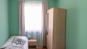 Ein kleines Zimmer mit zwei Betten, einem Holzschrank, einem Fenster mit durchsichtigen Vorhängen und einem Nachttisch mit Lampe.