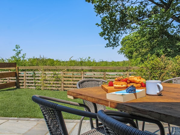 Un espace de restauration en plein air avec une table en bois, des chaises en osier et une vue sur une pelouse verdoyante et une clôture en bois.