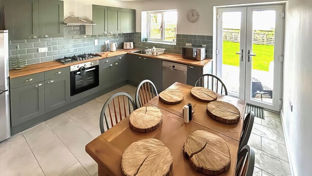 A modern kitchen with grey cabinets, a wooden dining table with a unique centerpiece, and a view of a green garden through the open door. - No. 4 Squirrels Nest-UK44305 (Great Witchingham)