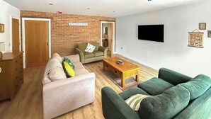 A living room with a brick wall, a green sofa, a pink sofa, a wooden coffee table, and a flat-screen TV mounted on the wall. - No. 4 Squirrels Nest-UK44305 (Great Witchingham)
